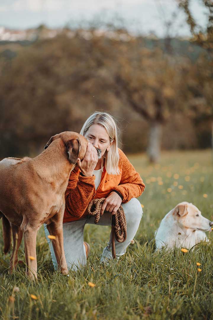 Juliane Dressel kniet auf einer Wiese und berührt sanft ihren Hund – ein Moment voller Vertrauen, Nähe und achtsamer Verbindung im Hundecoaching.