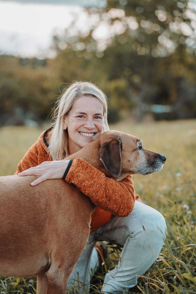 Juliane Dressel sitzt mit ihrem Hund auf einer Wiese und lächelt – sie begleitet Mensch und Hund auf dem Weg zu mehr Vertrauen und Verbindung.