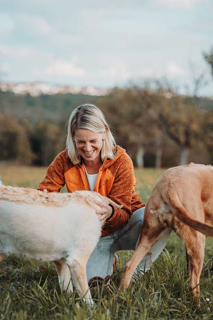 Juliane Dressel lacht herzlich, während sie mit ihren Hunden in einer Wiese spielt – Ausdruck von Leichtigkeit, Vertrauen und echter Verbindung im Hundecoaching.